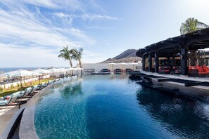 Un área de piscina con sillones de descanso y sombrillas, una zona de bar con asientos, y vista al océano y montañas al fondo.