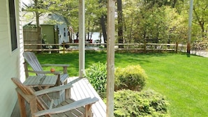 Une terrasse en bois avec deux fauteuils Adirondack, une petite table et un espace porche abrité donnant sur une cour verdoyante, avec une clôture en bois et un petit abri de jardin en arrière-plan.