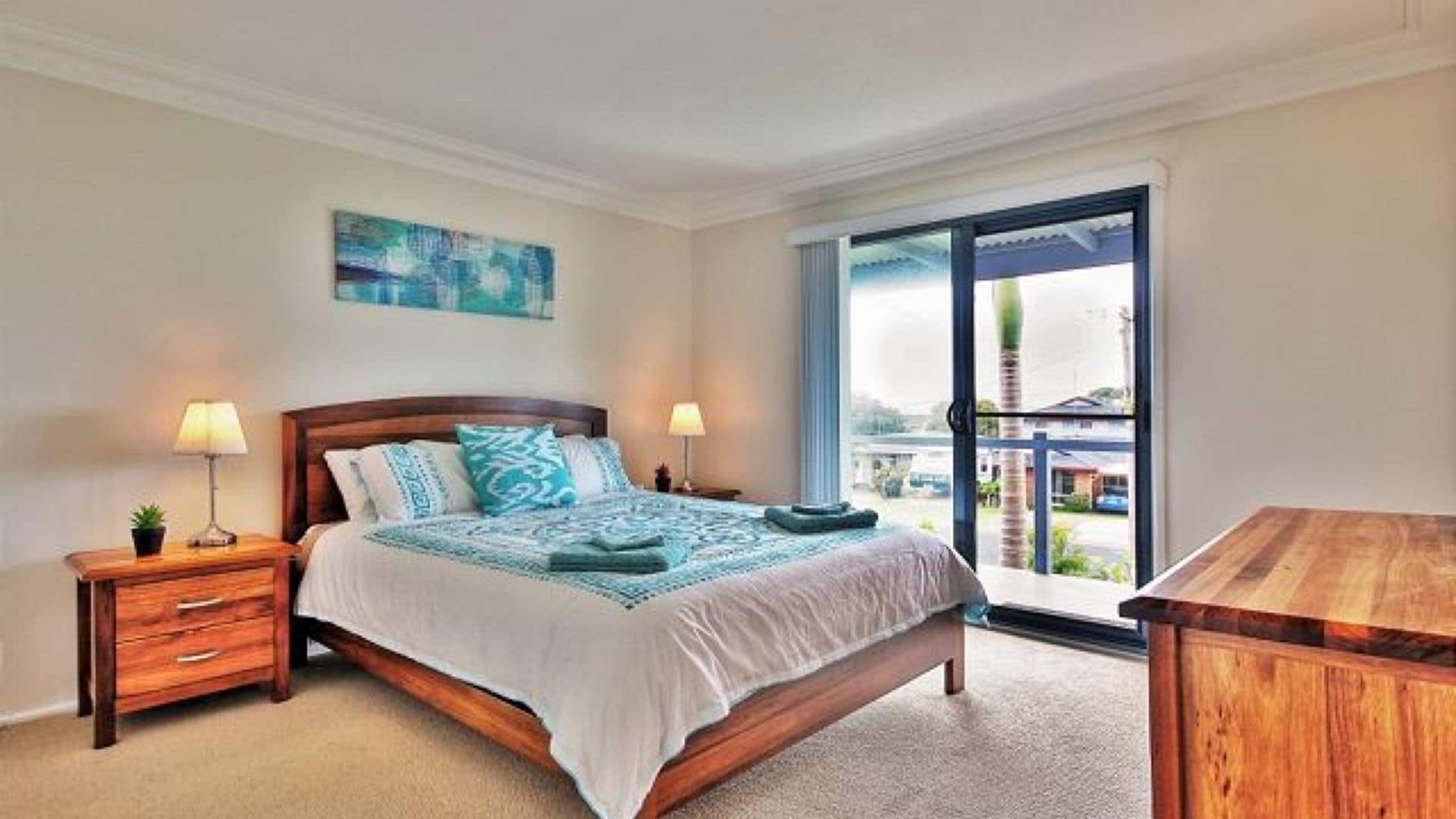 Um quarto com uma cama, duas mesas de cabeceira, uma mesa de apoio, uma escrivaninha de madeira, uma varanda com vista e um quadro na parede.