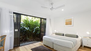 Un dormitorio con cama, mesita de noche, lámpara, ventilador de techo y vistas a zonas verdes a través de un ventanal corredero.