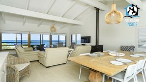 A spacious living area with a large wooden dining table, comfortable seating, a television, and a view of the ocean.