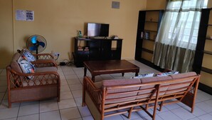 Ein Wohnzimmer mit einer Couch, einem Holztisch, einem Fernseher auf einem schwarzen Schrank, einem Ventilator und einem Bücherregal.
