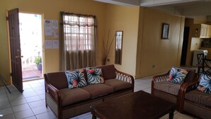 Ein Wohnzimmer mit Sofa, Sesseln und Couchtisch. Eine Tür führt nach draußen, ein Fenster mit Vorhängen und ein an der Wand befestigtes Schild.