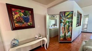Un salon avec un canapé blanc, une table basse en verre et un tableau représentant des fleurs tropicales. On y trouve également un coin cuisine avec des meubles blancs et une fenêtre équipée de stores.