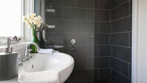 Ein modernes Badezimmer mit dunklen Fliesen, einem weißen Waschbecken, einem Spiegel und einer Vase mit Blumen.