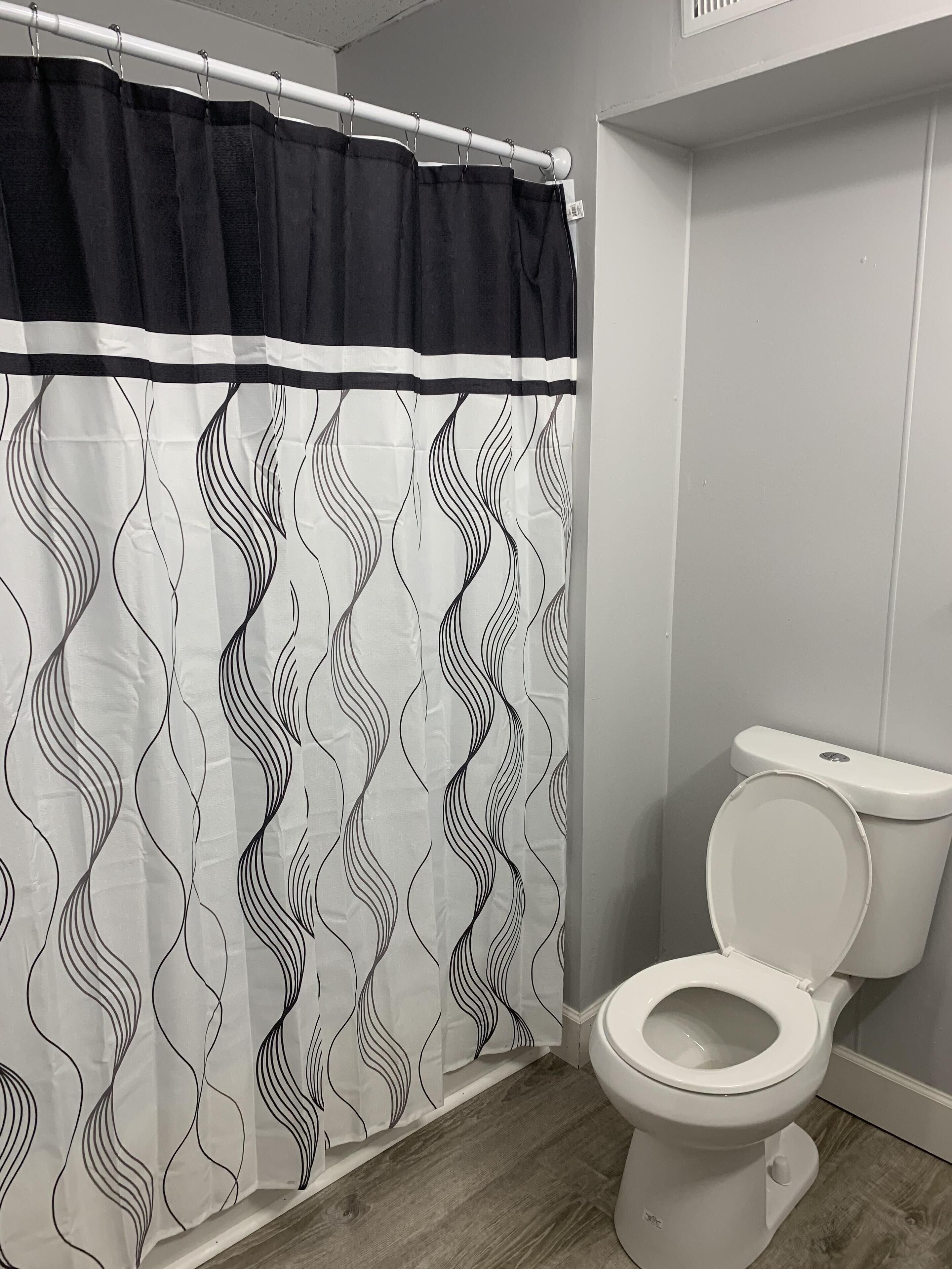Un baño con inodoro blanco, cortina de ducha de estampado blanco y negro, y piso de madera.