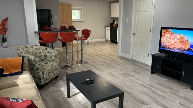 Una sala de estar moderna con un sofá, una mesa de centro y un televisor. Se observa una cocina al fondo con sillas rojas y una mesa de comedor.