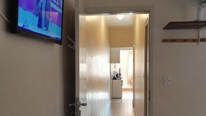 Un pasillo con una televisión mostrando 'Havan' en la pared, un ventilador montado y una cama con colchón blanco.