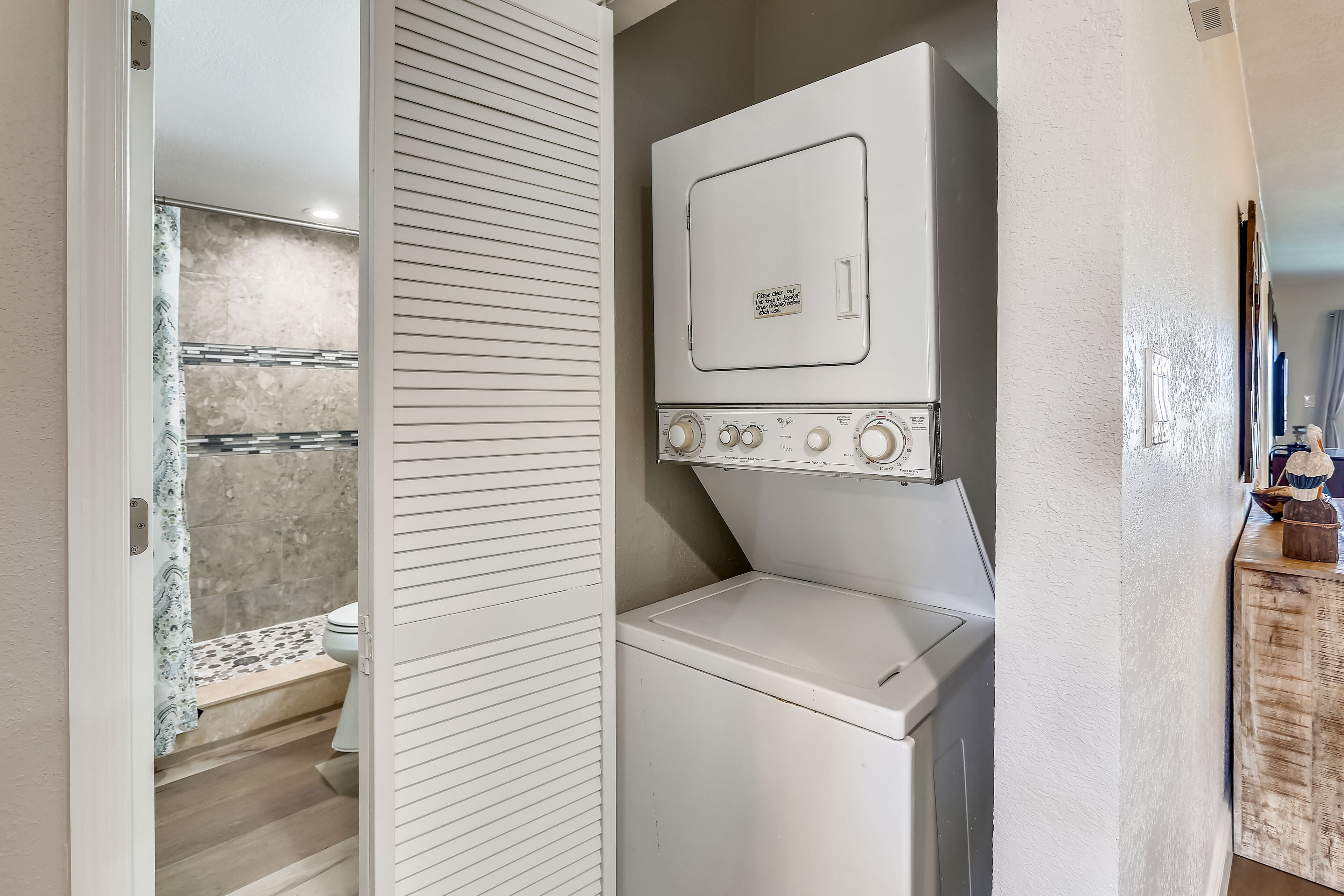 A compact laundry room with a front-loading washer and dryer, a wooden cabinet with decorative items, and a shower area with a patterned shower curtain.