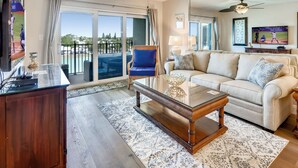 Un salón con sofá, mesa de centro y televisor. Tiene un balcón con vistas al exterior.