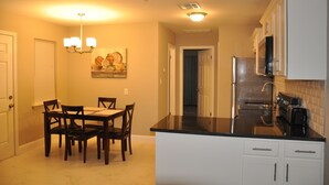 Een kleine keuken met witte kasten, een zwart aanrechtblad en een eethoek met een tafel en stoelen.