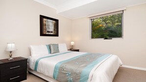Un dormitorio con cama, dos mesitas de noche, una mesilla auxiliar, un cuadro en la pared y una ventana con vistas a árboles.