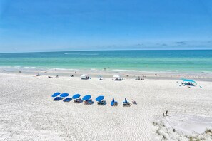 A beach with white sand and clear blue water. There are several blue umbrellas and lounge chairs arranged on the sand. - Villa Madeira 506 Amazing Updates|Great decor|Large Beach Front Balcony! (Madeira Beach)