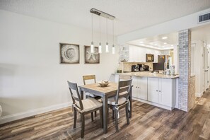 Un comedor con mesa y sillas de madera, una cocina con muebles blancos y lámparas colgantes.