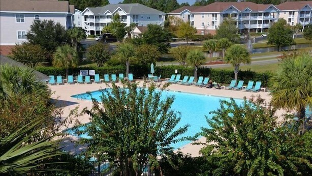 Ein Pool, umgeben von Liegestühlen und Palmen, mit Hotels im Hintergrund.