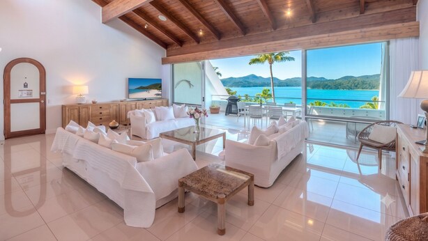 A spacious living room with white sofas, a glass coffee table, and a view of a beach and mountains through large windows. - Bella Vista East 9: 2 bedroom oceanview apartment with golf buggy (Hamilton Island)