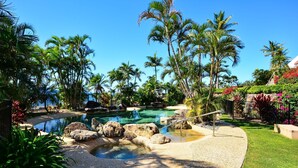A tropical resort with a large pool surrounded by palm trees and a small waterfall. - Bella Vista East 9: 2 bedroom oceanview apartment with golf buggy (Hamilton Island)