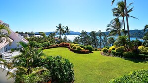 A well-maintained garden with manicured lawns, palm trees, and a view of a lake and mountains in the background. - Bella Vista East 9: 2 bedroom oceanview apartment with golf buggy (Hamilton Island)