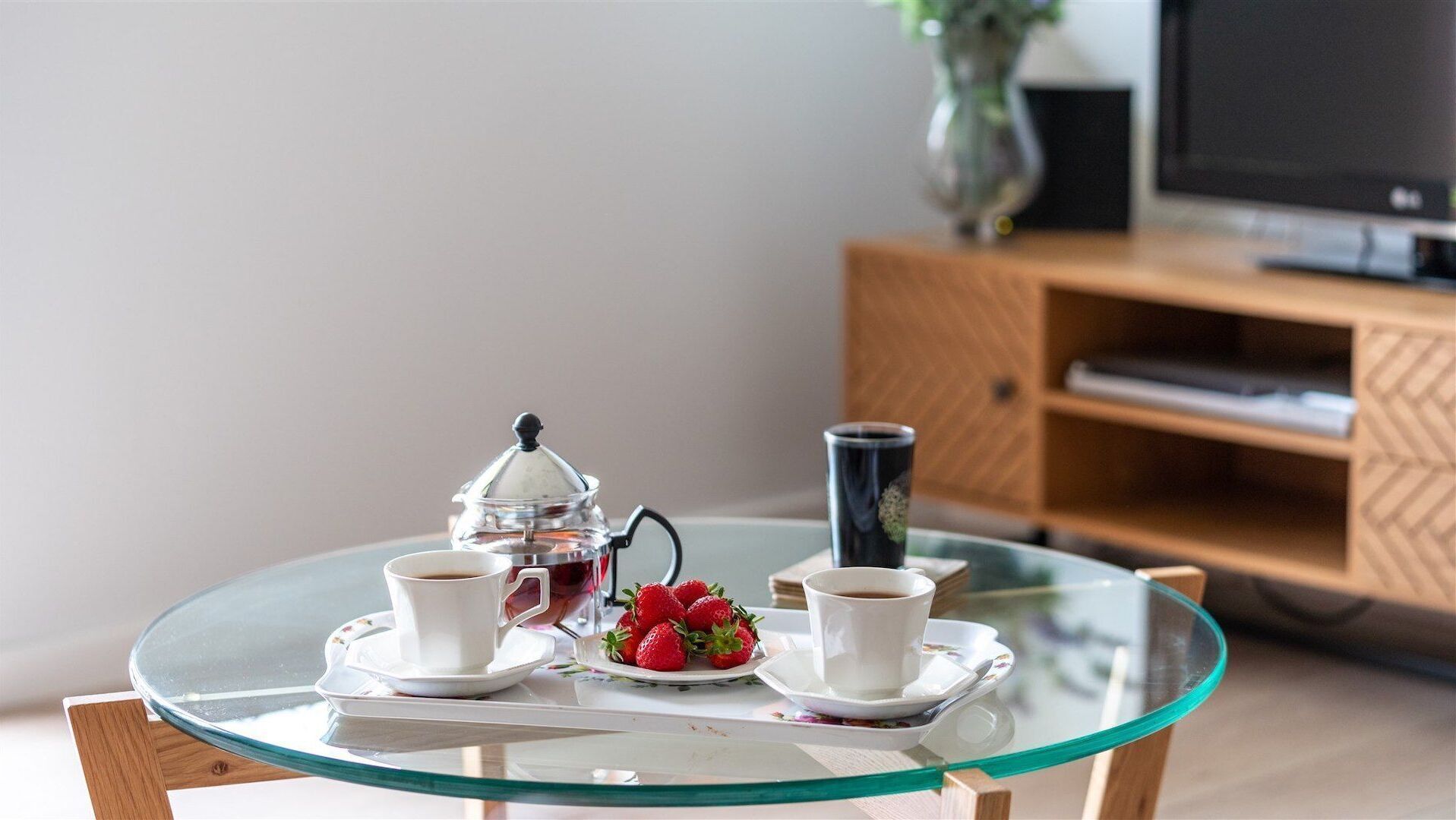 A glass table with a coffee pot, two cups, and a plate of strawberries.