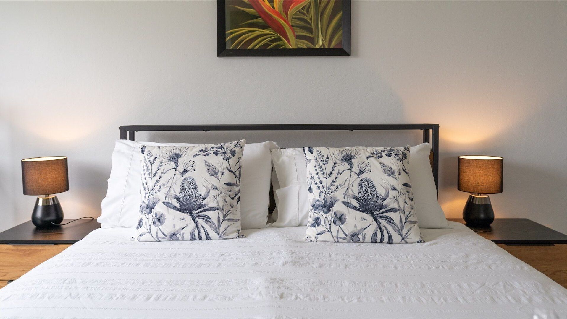 Una cama impecablemente hecha con dos almohadas de estampado floral, flanqueada por dos lámparas de noche sobre mesitas negras, frente a una pared lisa con una obra de arte enmarcada en la parte superior.