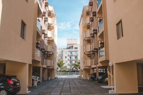 A narrow street between two multi-story buildings with balconies, parked cars, and a clear sky.