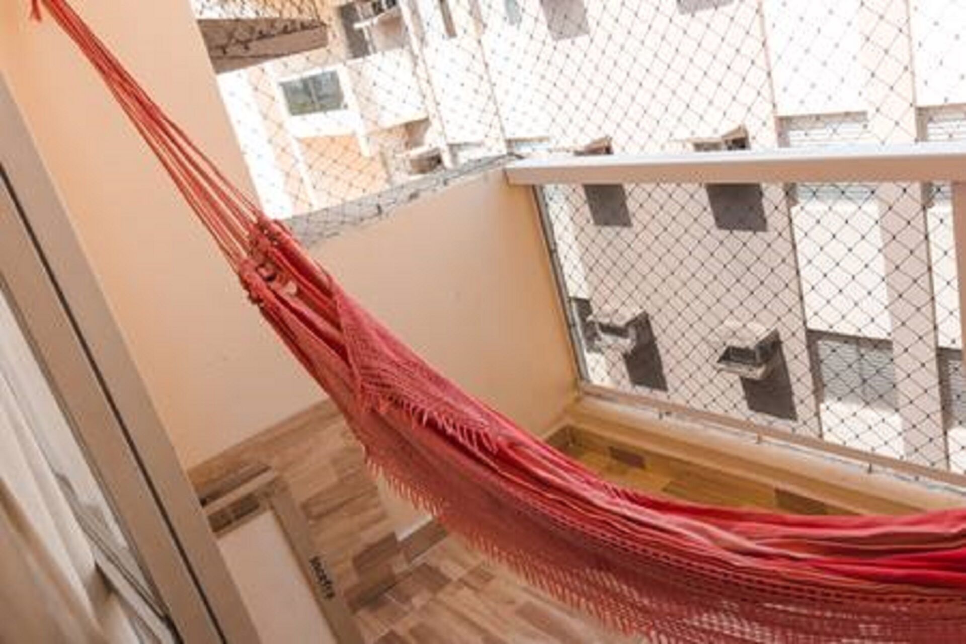 A red hammock hanging between a balcony and a window, with a view of buildings in the background.