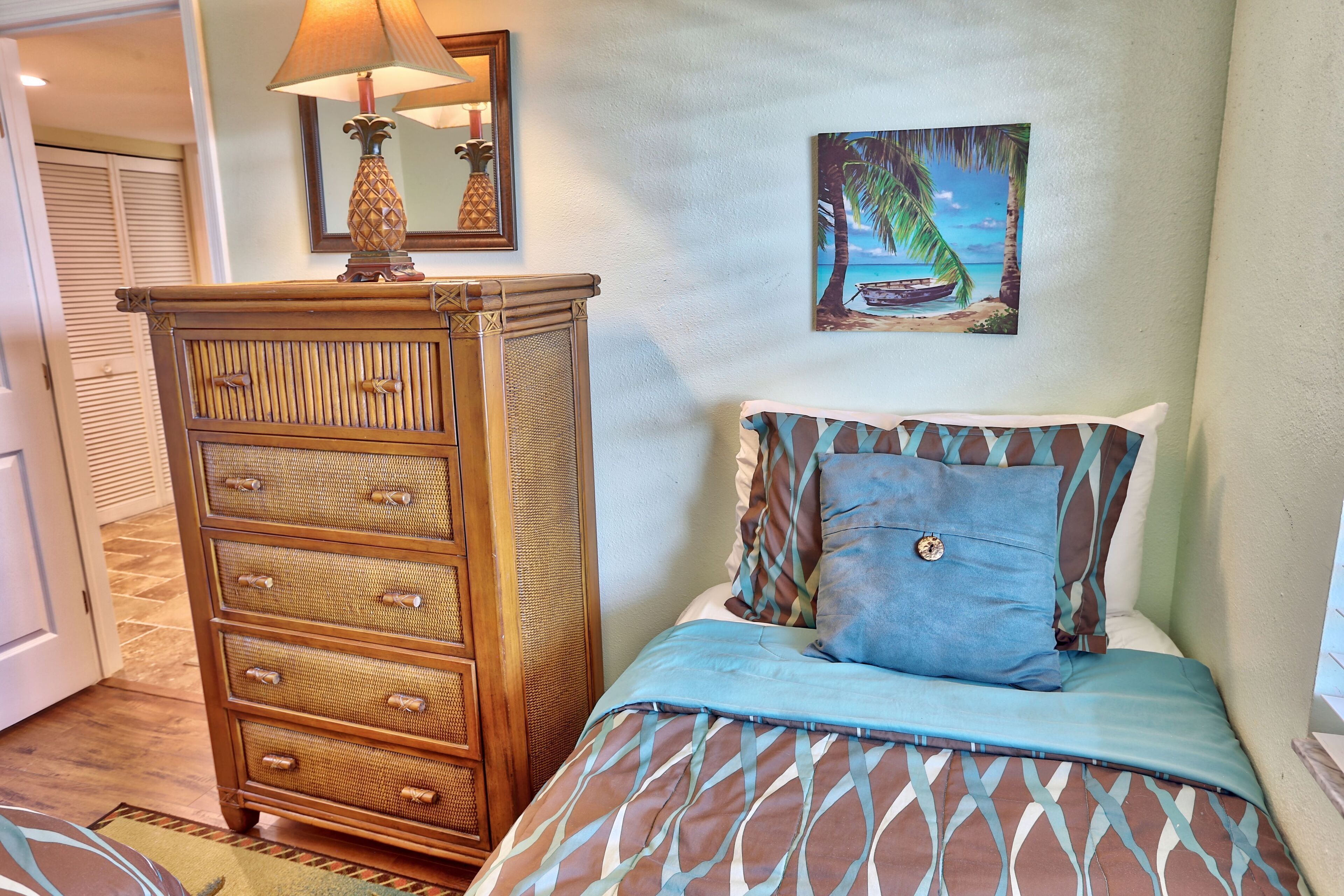 A bedroom with a bed, a dresser, a painting of a beach scene, and a lamp on the dresser.