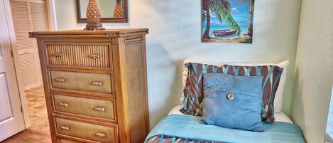 A bedroom with a bed, a dresser, a painting of a beach scene, and a lamp on the dresser.