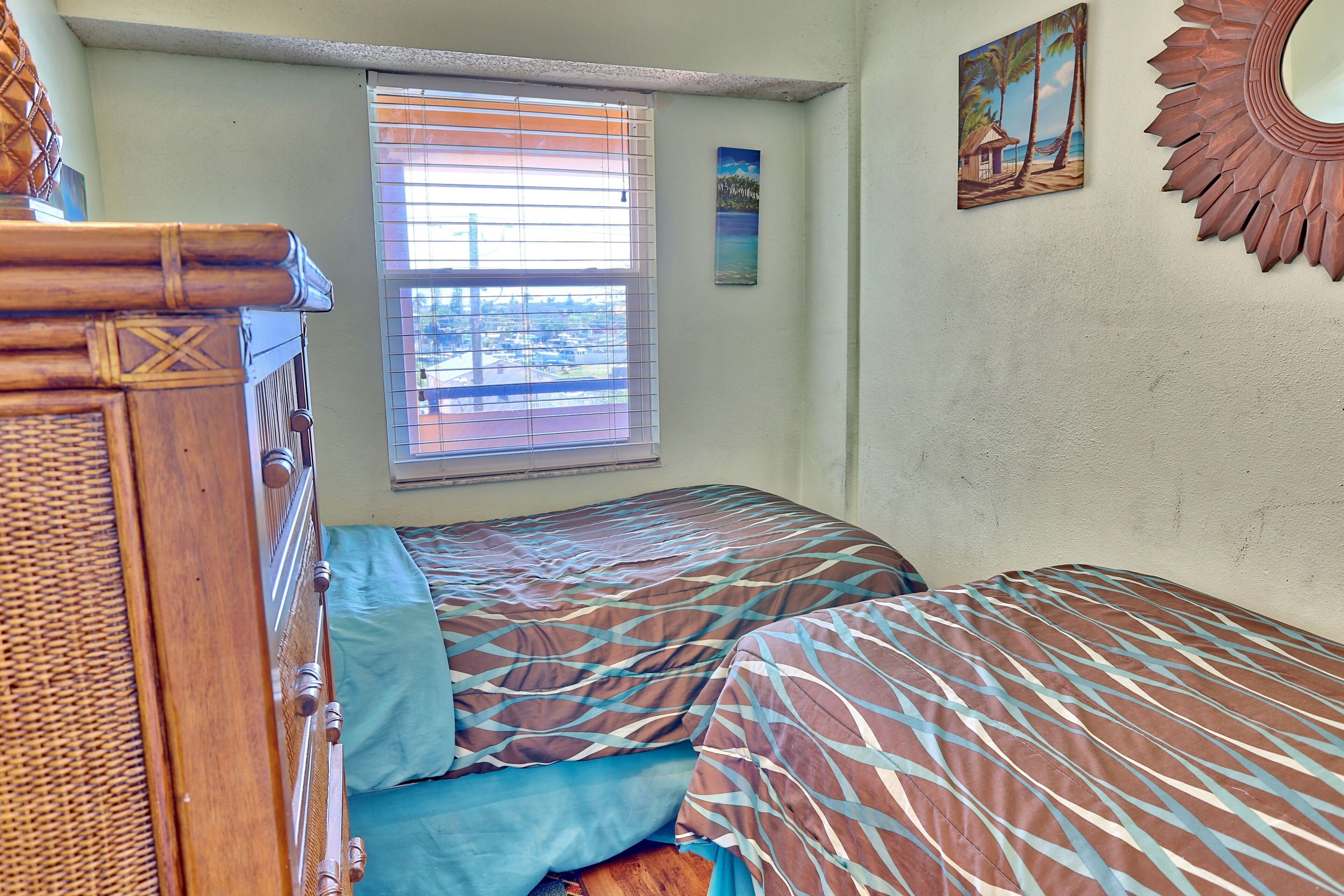 A bedroom with two beds, a window with blinds, and a wall-mounted mirror.