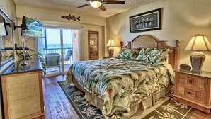 A bedroom with a bed, ceiling fan, two bedside lamps, a nightstand, a TV, and a view of the ocean through a sliding door.
