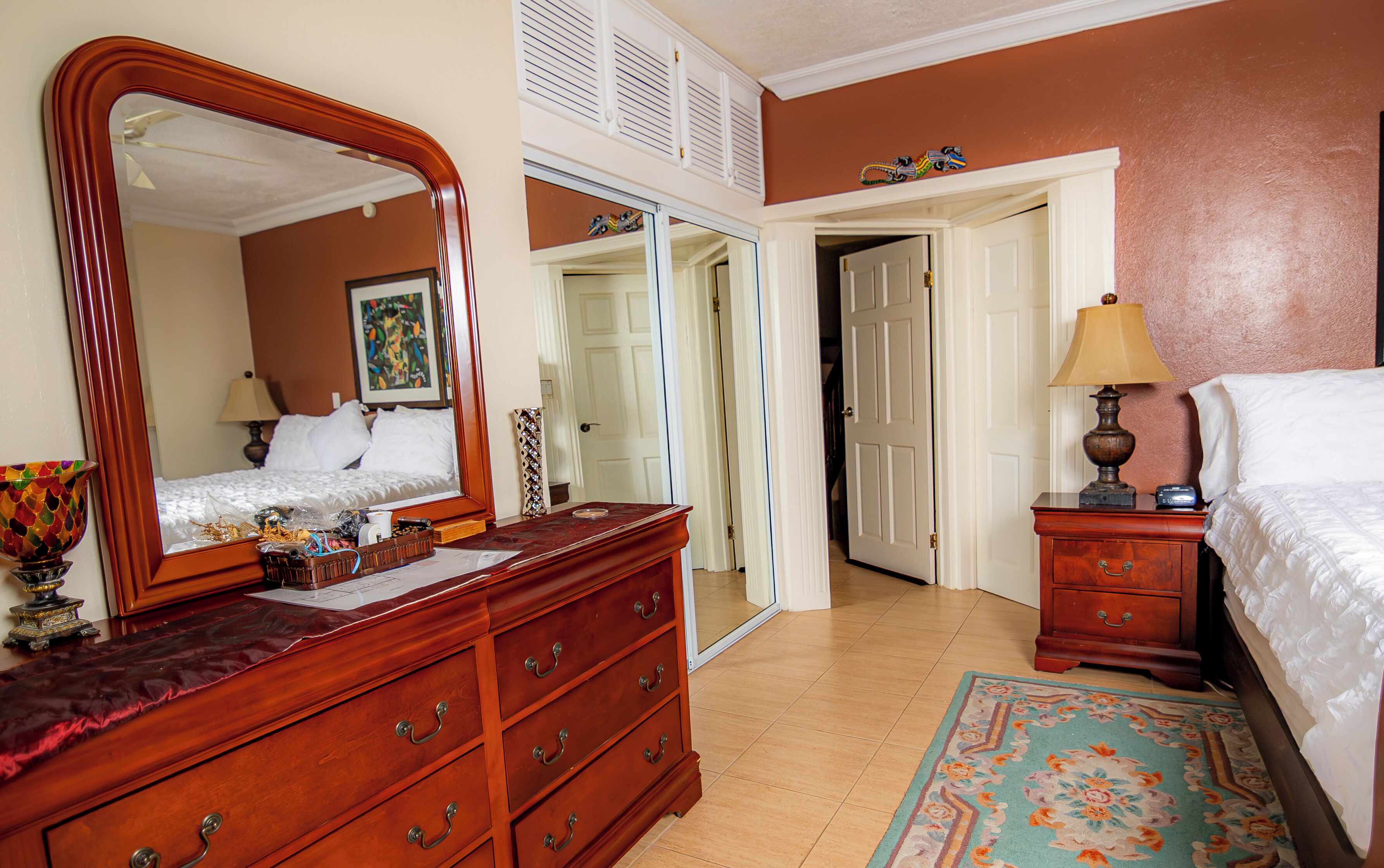 Une chambre d’hôtel avec une commode en bois, un grand miroir, un lit avec du linge de maison blanc, une table de chevet avec une lampe et une porte donnant sur un couloir.