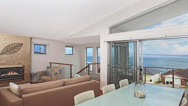 Amplia sala de estar con chimenea, un sofá y una mesa de comedor. Una gran puerta de vidrio da a un balcón con vistas al mar.