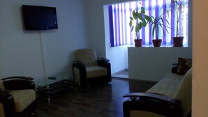 Een woonkamer met een flatscreen televisie aan de muur, een bank, een stoel, een bijzettafeltje en een raam met potplanten.