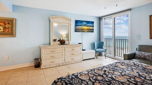 A bedroom with a large bed, a dresser, a chair, and a view of the ocean through large windows.