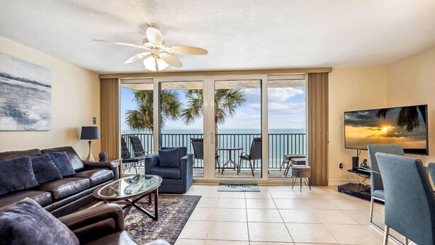 A living room with a sofa, coffee table, and TV, a balcony with chairs, and a view of the ocean.