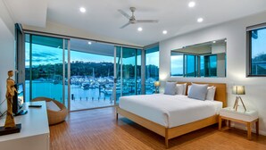 A modern bedroom with a large bed, a desk with a computer, and a view of a marina through floor-to-ceiling glass doors. - Pavillions Penthouse 25 - Waterfront with private pool, golf buggy and transfers (Hamilton Island)