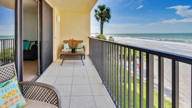 Un balcón con vista a la playa, una silla de mimbre, una mesa con un florero y una palmera.