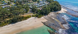 An aerial view of a coastal area with a cluster of houses near the beach, surrounded by greenery and rocky coastline. - Bombara by Experience Jervis Bay (Huskisson)