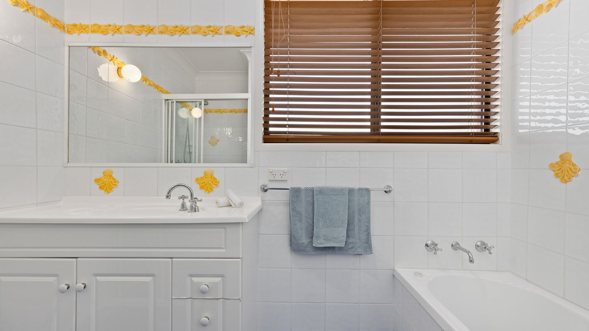 Un baño con azulejos blancos, un lavabo con espejo, una ventana con persianas y una bañera.