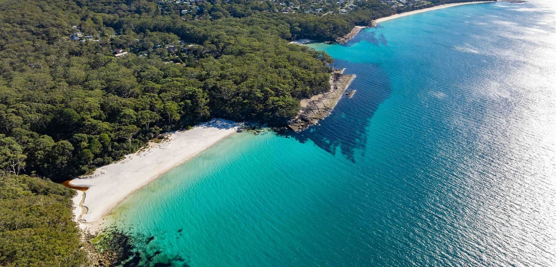 An aerial view of Jervis Bay with a beach, clear turquoise waters, and dense forested areas.