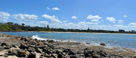 A rocky beach with clear blue water and a distant shoreline with buildings and trees.