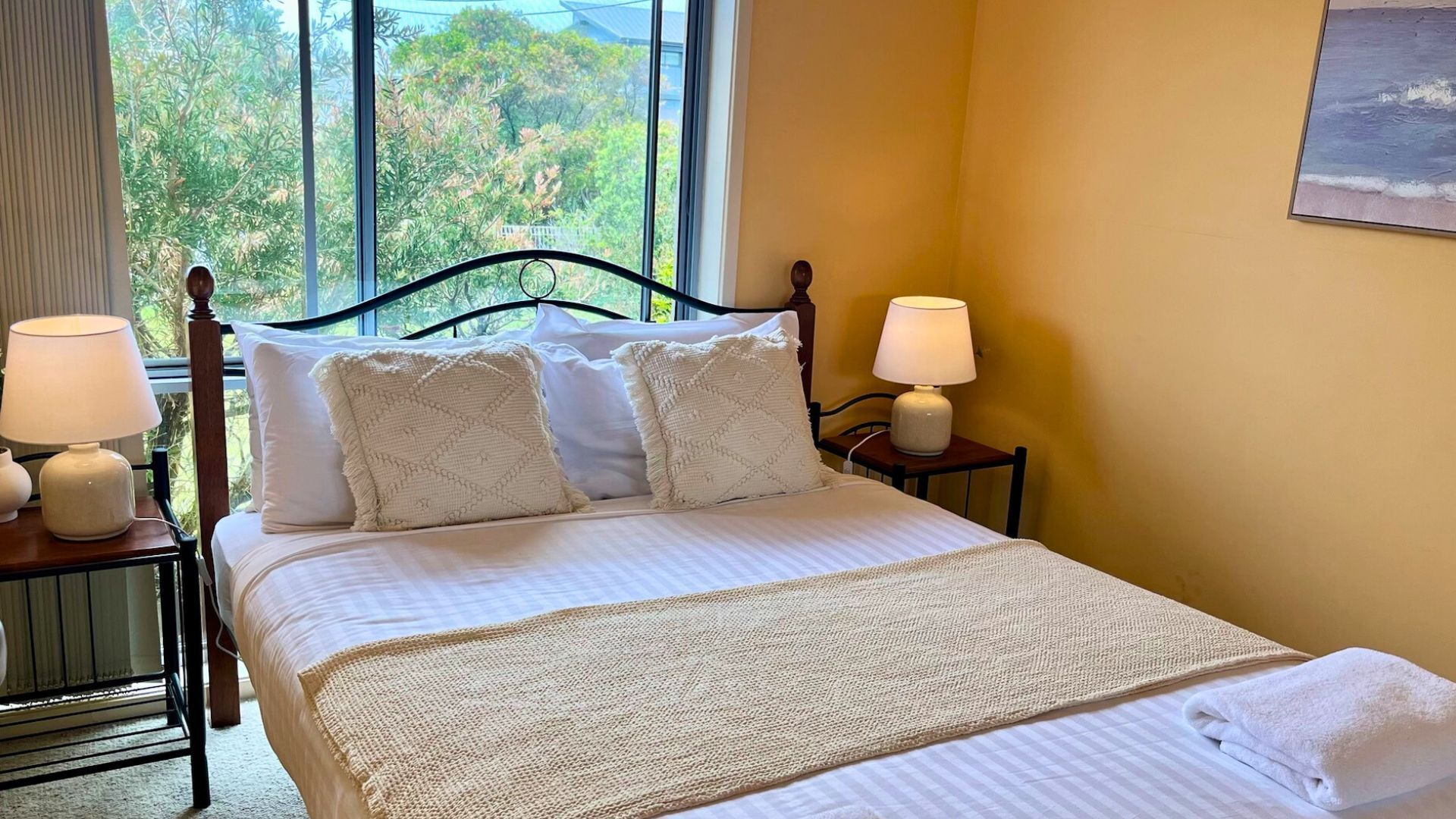 A neatly made bed with a white comforter and two decorative pillows, flanked by two bedside tables with lamps, in a room with a large window offering a view of greenery outside.