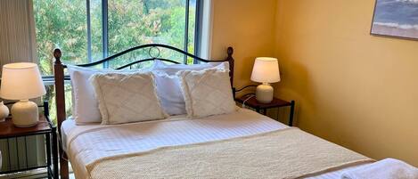 A neatly made bed with a white comforter and two decorative pillows, flanked by two bedside tables with lamps, in a room with a large window offering a view of greenery outside.