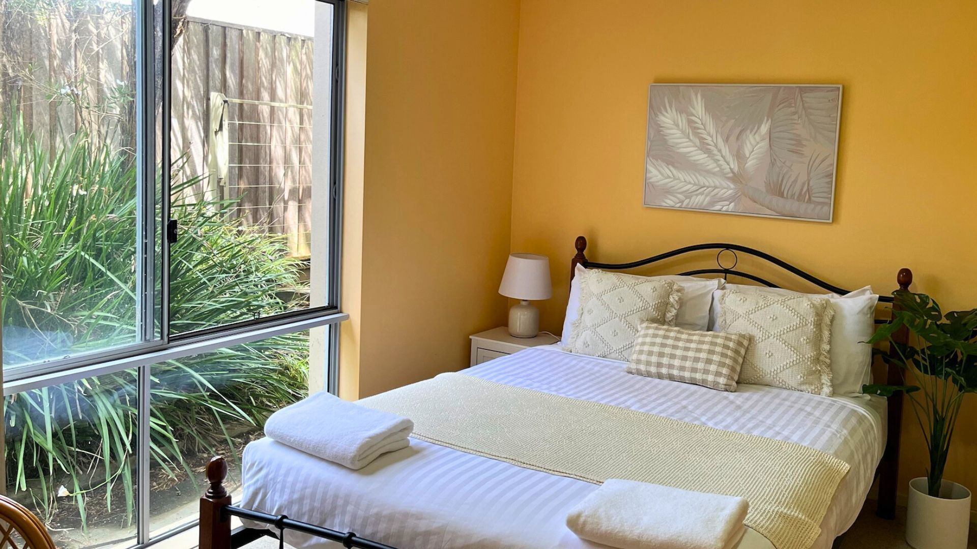 Un dormitorio con un ventanal grande, una cama con ropa blanca, una mesita de noche con lámpara y una obra de arte colgada en la pared.