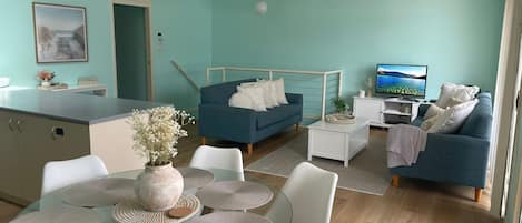 Una sala de estar moderna con un sofá azul, un televisor y un comedor con mesa de vidrio.
