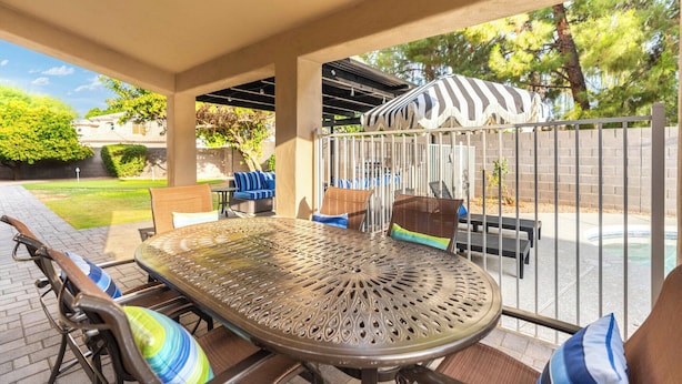 Uma área de varanda coberta com mesa e cadeiras de metal, um guarda-sol listrado e vista para uma piscina.