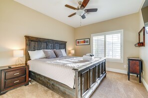 Une chambre avec un lit en bois, un ventilateur de plafond, une fenêtre avec des stores, une table de chevet avec une lampe et un tableau encadré au mur.
