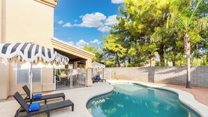 Piscina com cadeiras de espreguiçadeira e guarda-sóis, cercada por um muro de tijolos e árvores.