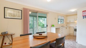 A dining area with a wooden table and chairs, a kitchen with white cabinets, and a window with a view of greenery. - Beachwood  on  Casey (Mystery Bay)
