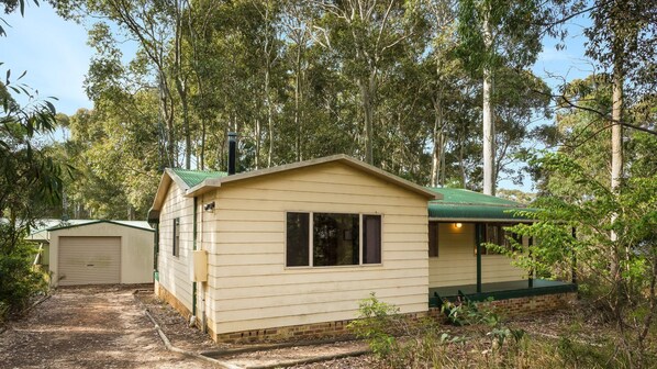 A small, single-story wooden cabin with a green roof, surrounded by dense forest.